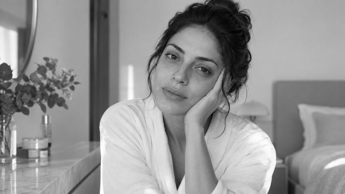 Mulher de roupão branco apoia o rosto na mão, com expressão cansada, sentada ao lado da cama num quarto iluminado
